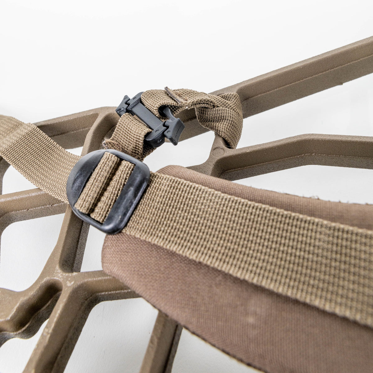 Close-up of a tan webbed strap with a black plastic buckle attached to a brown padded item, shown against a white background.